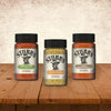 Three bottles of Stubb's seasoning including Garlic Lime, Texas Smokehouse Style, and the center bottle of Stubbs Hkry Must E are displayed on a wooden table against a textured wall.