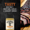 A bottle of Stubbs Hkry Must E seasoning is displayed against a dark textured background next to a sliced smoked brisket to highlight its flavors of sea salt mustard garlic and hickory smoke.
