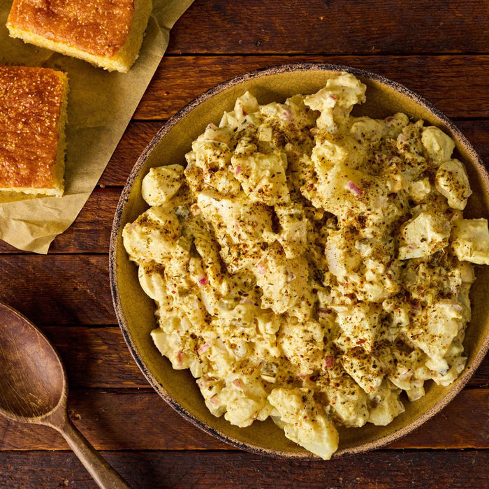 A rustic bowl of creamy potato salad seasoned with Stubbs Hkry Must E sits on a wooden table alongside pieces of golden cornbread.