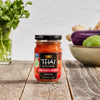 A jar of Thai Kitchen red curry paste sits on a wooden table with vegetables in the background.