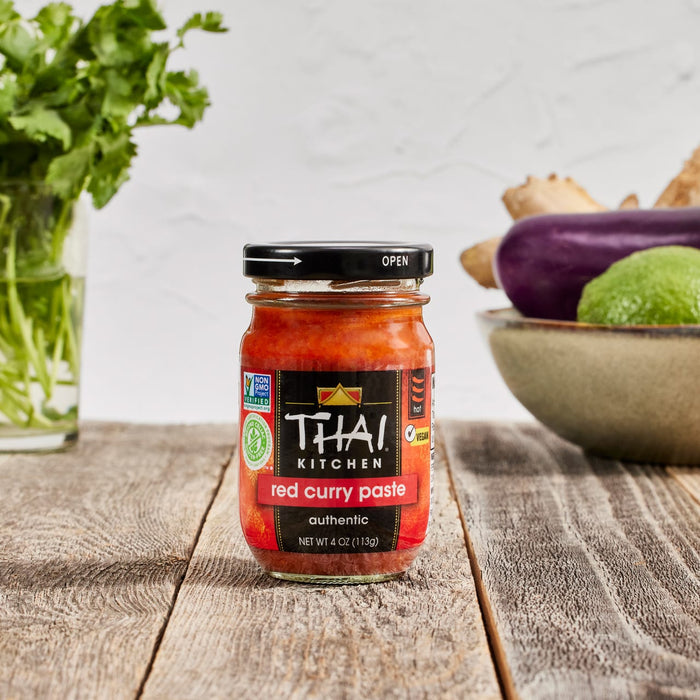 A jar of Thai Kitchen red curry paste sits on a wooden table with vegetables in the background.