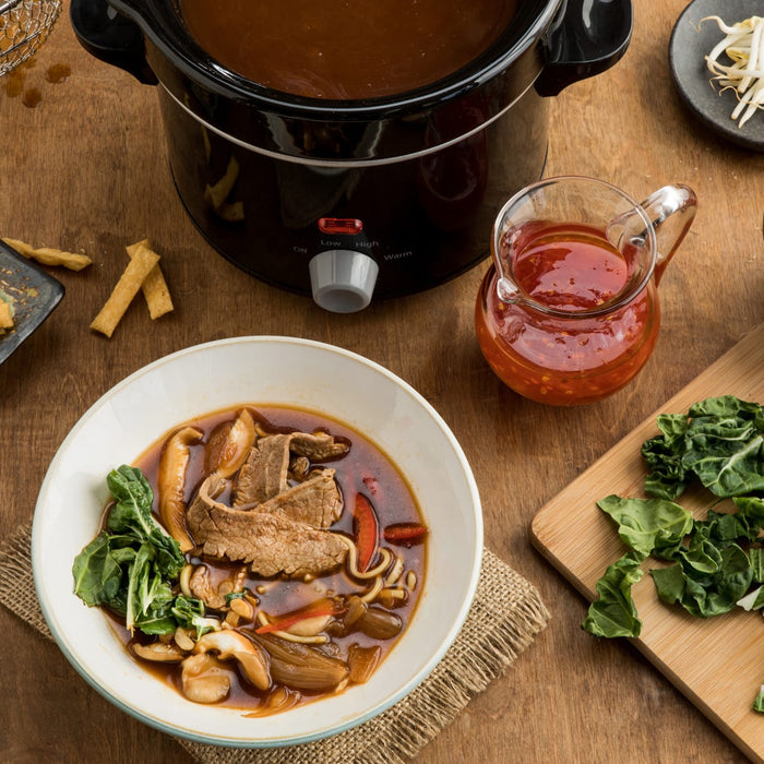 A bowl of beef noodle soup with a side of greens and a small pitcher of red sauce.
