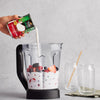 A hand pours So Delicious coconut milk into a blender containing berries, preparing a smoothie.