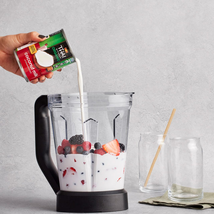 A hand pours So Delicious coconut milk into a blender containing berries, preparing a smoothie.