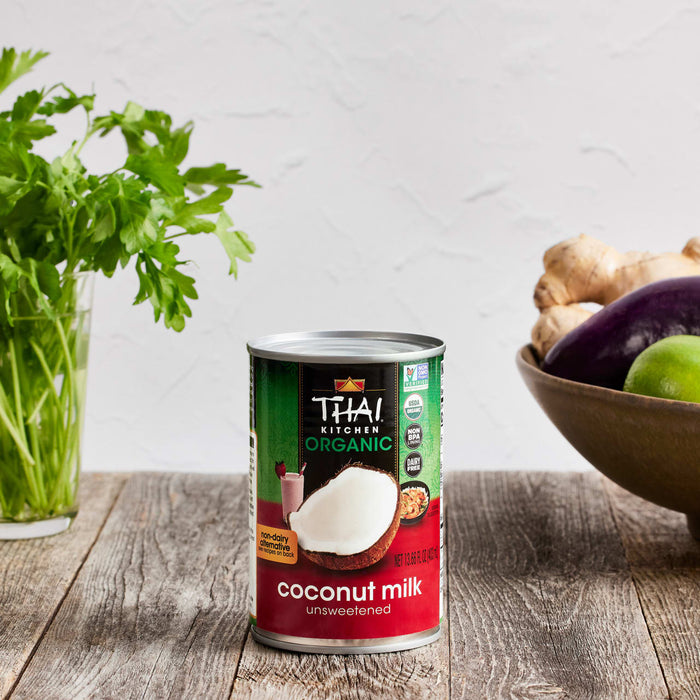 A can of Thai Kitchen Organic Coconut Milk sits on a wooden surface, surrounded by fresh ingredients like ginger and eggplant.