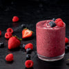 A vibrant berry smoothie is displayed in a glass, surrounded by fresh strawberries, raspberries, and blueberries on a dark surface.