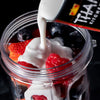 A close-up of a blender with coconut milk being poured over strawberries raspberries and blueberries.