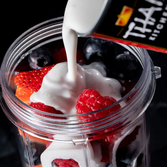 A close-up of a blender with coconut milk being poured over strawberries raspberries and blueberries.