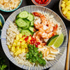 A bowl of rice with shrimp, mango, cucumber, coconut flakes, lime wedges, and cilantro.