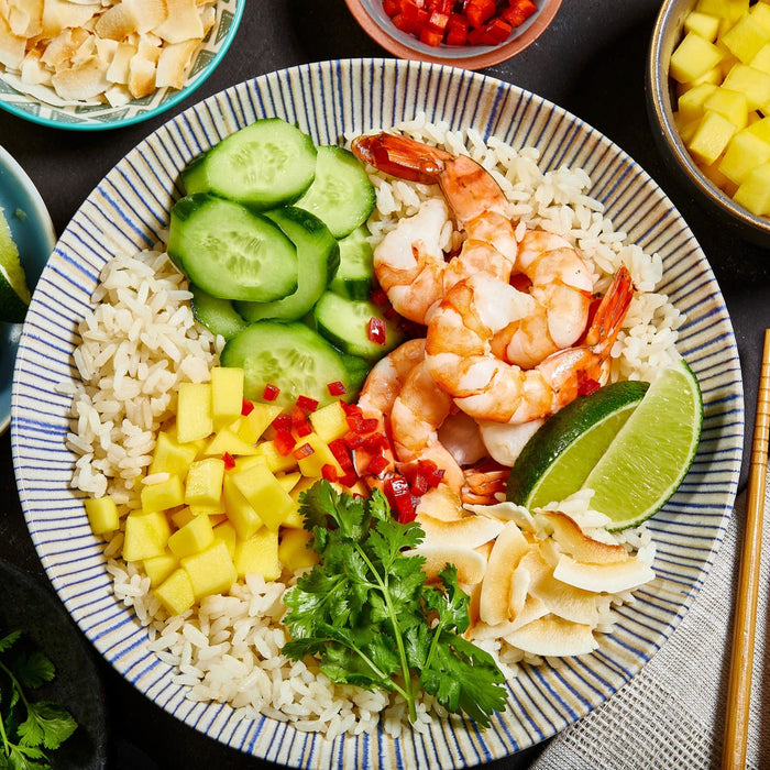A bowl of rice with shrimp, mango, cucumber, coconut flakes, lime wedges, and cilantro.