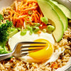 A fork is placed on a bowl of rice with a fried egg, avocado, broccoli, carrots, and green onions.