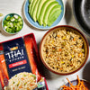 A package of Thai Kitchen fried rice next to a bowl of the prepared rice, a bowl of sliced avocado, a bowl of chopped green onions, and a bowl of shredded carrots.