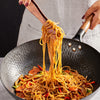 A person is using wooden tongs to toss noodles in a wok.
