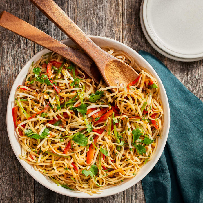 A bowl of noodles with vegetables and peanuts with two wooden spoons on top.