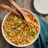 A bowl of noodles with vegetables and peanuts with two wooden spoons on top.