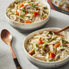 Two bowls of chicken noodle soup with mushrooms and carrots are displayed next to a wooden spoon and a product box.