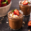 Two jars of chocolate pudding are garnished with strawberries and coconut flakes, with a bowl of fresh strawberries in the background.