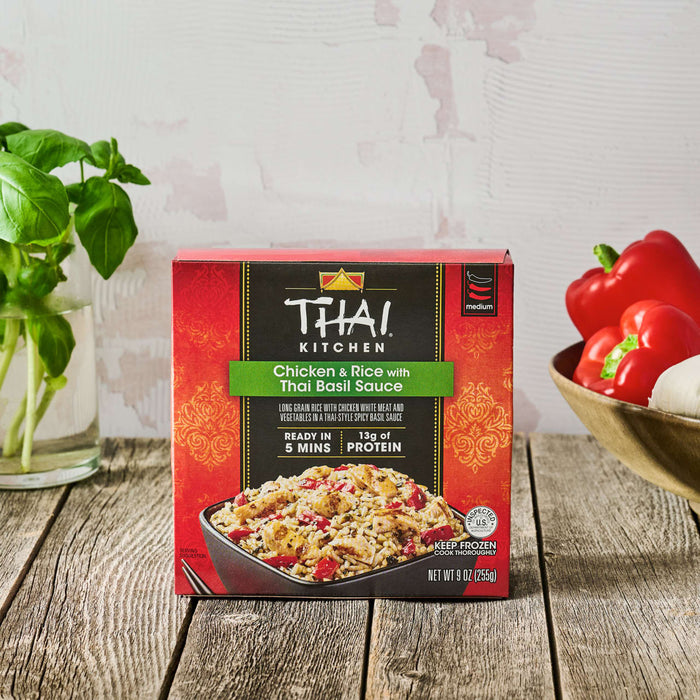 A box of Thai Kitchen, 9.0 Oz Chicken &amp; Rice with Thai Basil Sauce is set on a rustic wooden table, surrounded by fresh ingredients like basil, red bell peppers, and garlic.