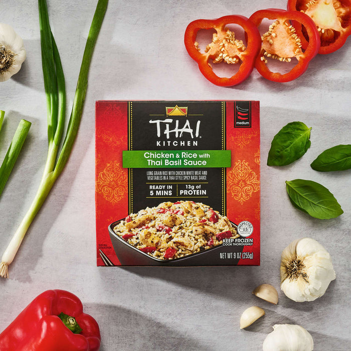 A box of Thai Kitchen, 9.0 Oz Chicken &amp; Rice with Thai Basil Sauce is displayed on a gray surface, surrounded by fresh ingredients like red bell peppers, garlic, scallions, and basil leaves.