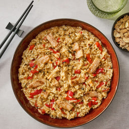 A top-down view of a creamy chicken and rice dish with red peppers, prepared using Thai Kitchen, 9.0 Oz, served in a brown bowl with chopsticks, a drink, and nuts on the side.