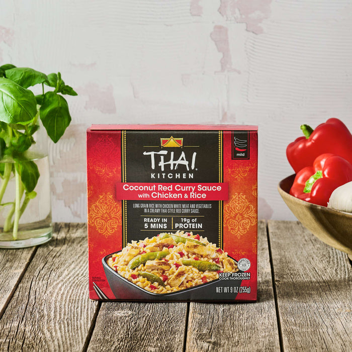 A box of Thai Kitchen, 9.0 Oz Coconut Red Curry Sauce with Chicken &amp; Rice sits on a rustic wooden table, surrounded by fresh ingredients like basil and red peppers.
