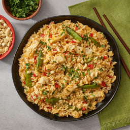 A black bowl filled with a finished recipe of chicken and rice made with Thai Kitchen, 9.0 Oz, is shown with side bowls of peanuts and cilantro.