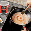 A person is stirring Thai Kitchen red curry sauce into a pan of coconut milk on a stovetop, with a container of the sauce visible in the background.