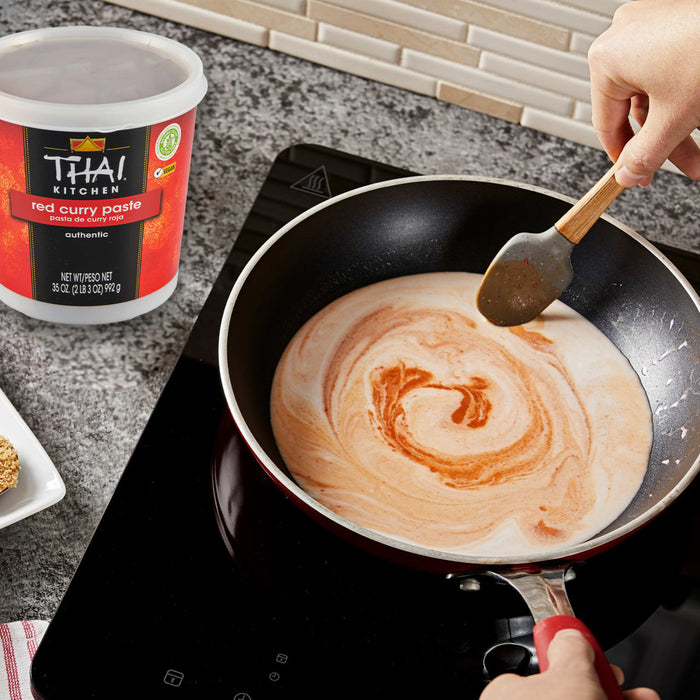 A person is stirring Thai Kitchen red curry sauce into a pan of coconut milk on a stovetop, with a container of the sauce visible in the background.