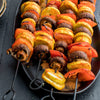 Grilled vegetable skewers featuring mushrooms, zucchini, and red bell peppers are arranged on a black platter.