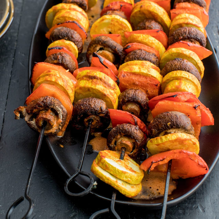 Grilled vegetable skewers featuring mushrooms, zucchini, and red bell peppers are arranged on a black platter.