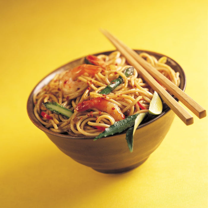 A bowl of noodles with shrimp, asparagus, and lime wedges, with chopsticks resting on the rim.