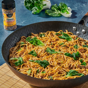 A wok filled with stir-fried noodles and bok choy, with a bottle of McCormick Montreal Steak Spice seasoning visible in the background.