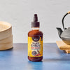 A bottle of Trader Joe's Black Garlic Hot Sauce stands on a blue surface, with a steamer and teapot in the background, highlighting its Asian-inspired flavor profile.