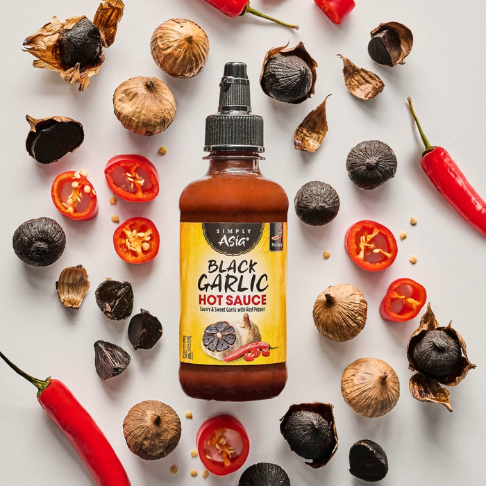 A bottle of Trader Joe's Black Garlic Hot Sauce is surrounded by black garlic cloves, red chili peppers, and halved cherry tomatoes.