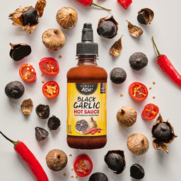 A bottle of Trader Joe's Black Garlic Hot Sauce is surrounded by black garlic cloves, red chili peppers, and halved cherry tomatoes.