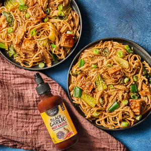 Two bowls of stir-fried noodles with vegetables and tofu are displayed next to a bottle of Yellowbird Habanero Condiment.