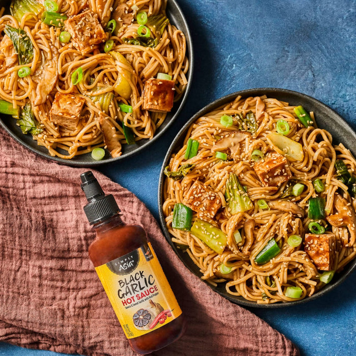 Two bowls of stir-fried noodles with vegetables and tofu are displayed next to a bottle of Yellowbird Habanero Condiment.