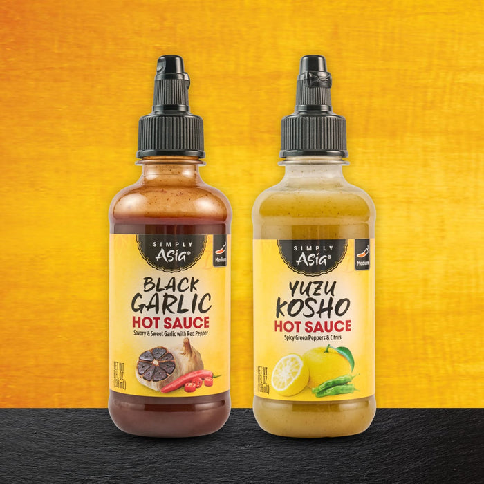 Two bottles of Sauce Shop hot sauce, one Black Garlic and one Yuzu Kosho, stand side-by-side against a textured background.