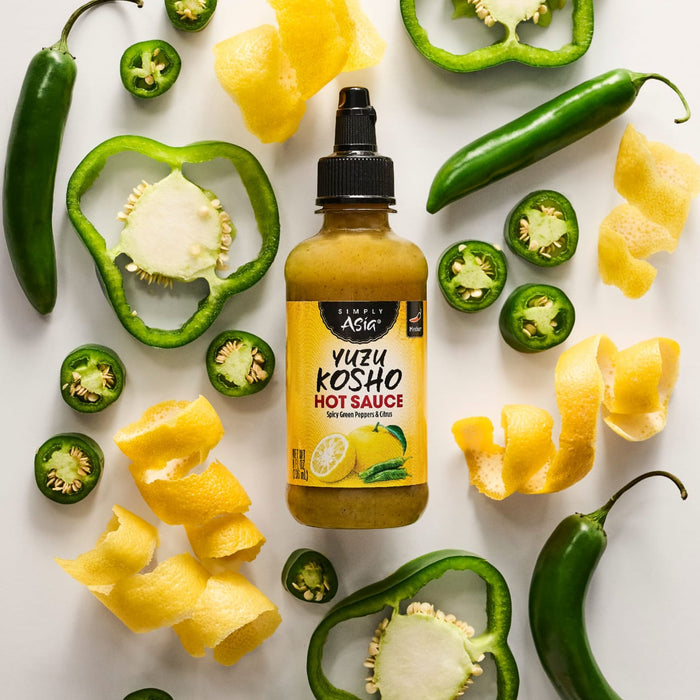 A bottle of Yellowbird Yuzu Hot Sauce is surrounded by fresh jalapeA?os, green bell pepper slices, and lemon zest.