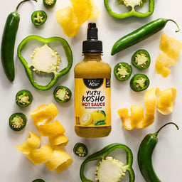 A bottle of Yellowbird Yuzu Hot Sauce is surrounded by fresh jalapeA?os, green bell pepper slices, and lemon zest.