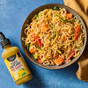 A bowl of stir-fried noodles with vegetables is displayed next to a bottle of Yellowbird Yuzu Kosho Hot Sauce.