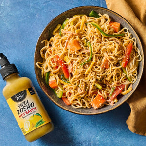 A bowl of stir-fried noodles with vegetables is displayed next to a bottle of Yellowbird Yuzu Kosho Hot Sauce.