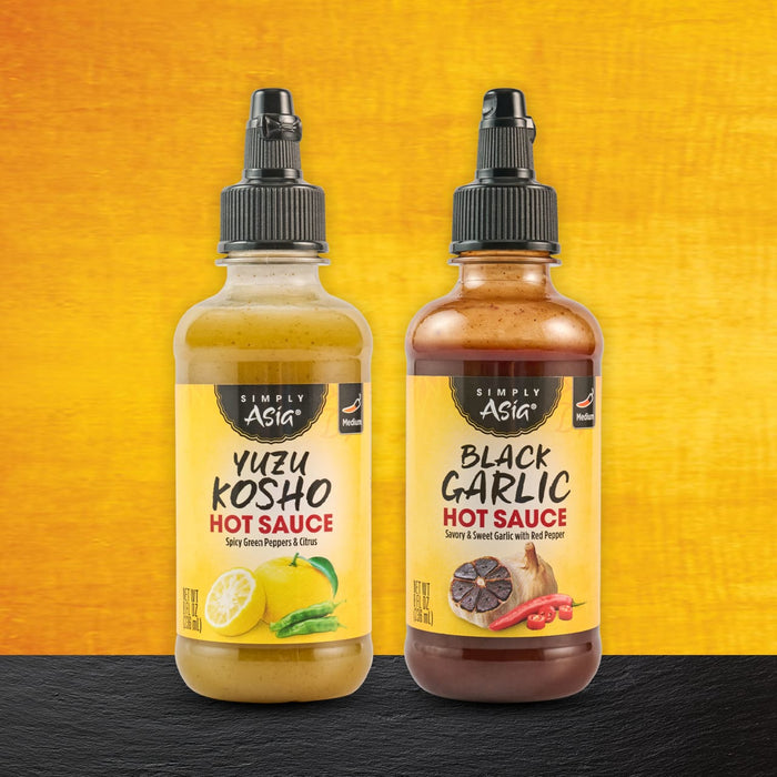 Two bottles of Master Sauce hot sauce, one Yuzu Kosho and one Black Garlic, are displayed against a vibrant background.