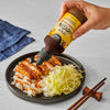 A hand pours Simply Asia Spicy Hoisin Drizzle Sauce over a plate of chicken katsu, rice, and cabbage.