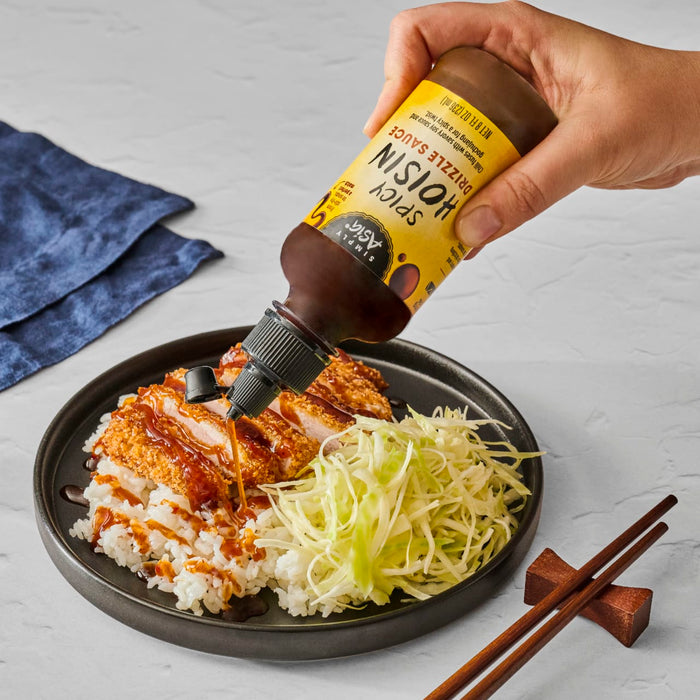 A hand pours Simply Asia Spicy Hoisin Drizzle Sauce over a plate of chicken katsu, rice, and cabbage.