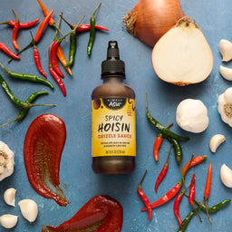 A bottle of Simply Asia Spicy Hoisin Drizzle Sauce is surrounded by red and green chili peppers, garlic cloves, and an onion on a blue surface.