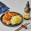 A plate of food featuring sliced, fried meat over rice and shredded cabbage, drizzled with Simply Asia Spicy Hoisin Drizzle Sauce, next to a bottle of the sauce and chopsticks.