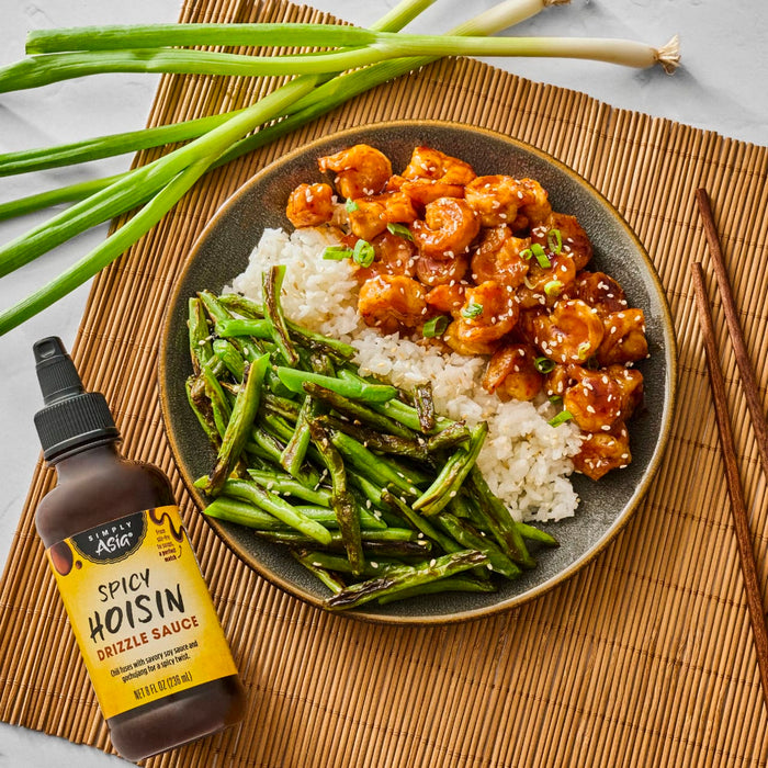 A bowl of rice, green beans, and spicy hoisin shrimp is displayed next to a bottle of Simply Asia Spicy Hoisin Drizzle Sauce.