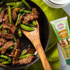 A close-up of a pan of beef and snow peas with a wooden spoon and a tube of Gourmet Garden Ginger Paste.