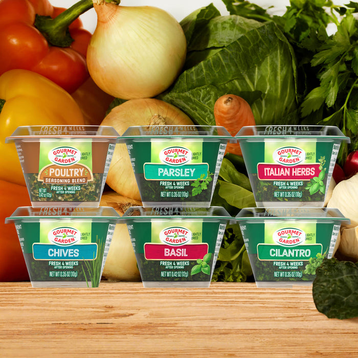 Six Gourmet Garden lightly dried herb and seasoning containers, including Gourmet Garden Basil (Case Of 72), are arranged on a wooden surface with fresh vegetables like peppers, onions, and cabbage in the background.